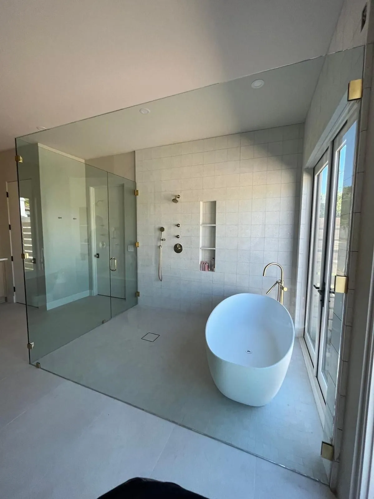 Modern bathroom with glass shower enclosure, freestanding tub, and window; white and gold accents.